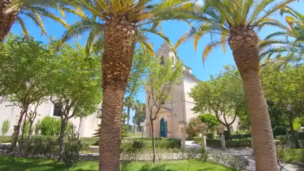 Two tall palm trunks framing a pale stone church with a bell tower, set among trimmed hedges and mixed garden trees.