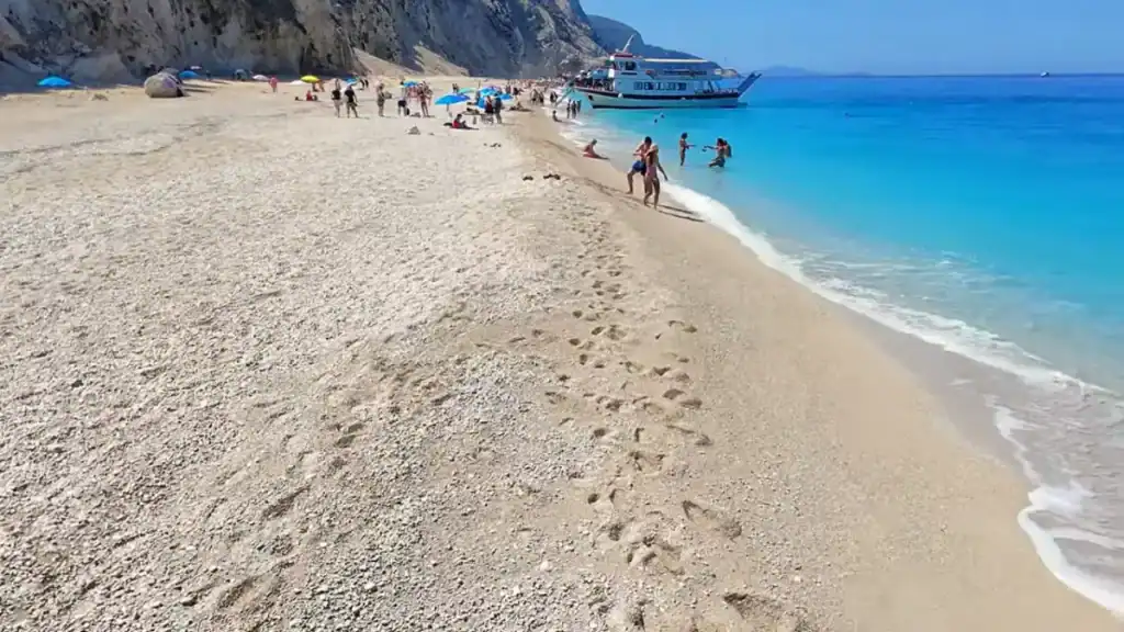 Coarse pebble and gravel beach surface in the foreground, a large weathered rock outcrop to the right, beachgoers with umbrellas
clustered toward the water's edge further along.