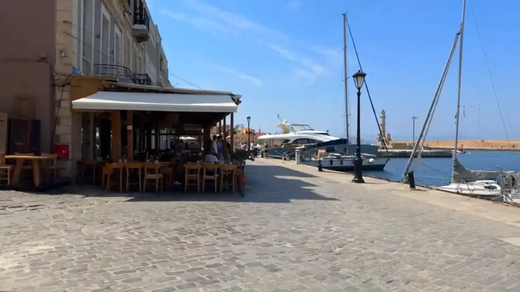 Cobbled quayside at Chania old harbour with a waterfront cafe, moored sailboats, and the Egyptian lighthouse visible at the harbour mouth