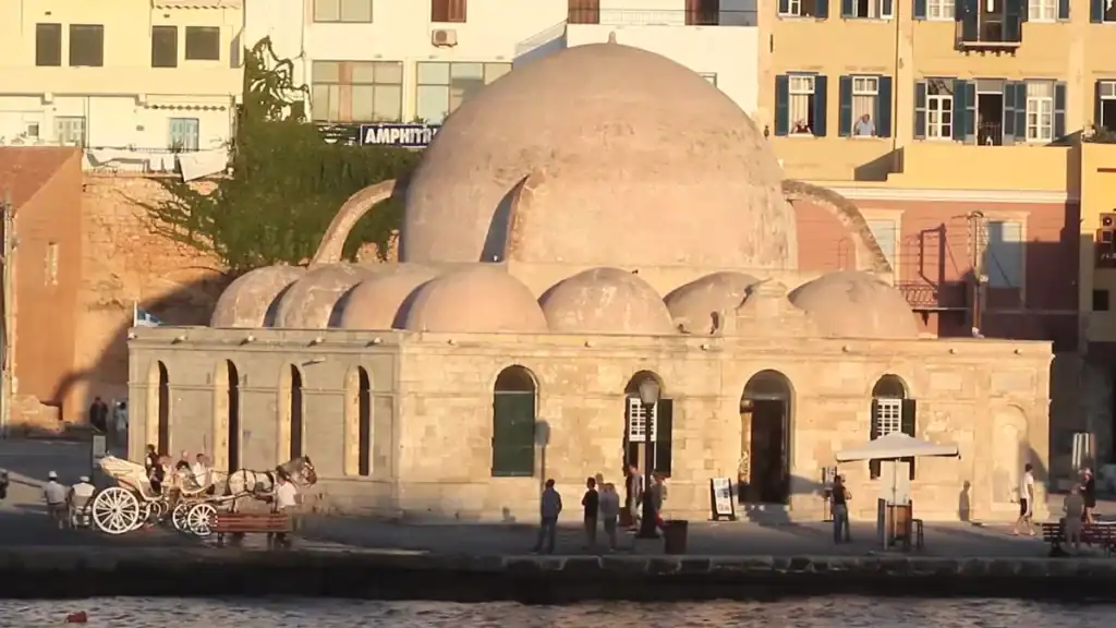 Ottoman domed mosque with clustered smaller domes and arched windows standing at the harbour edge, pedestrians and a horse-drawn carriage nearby