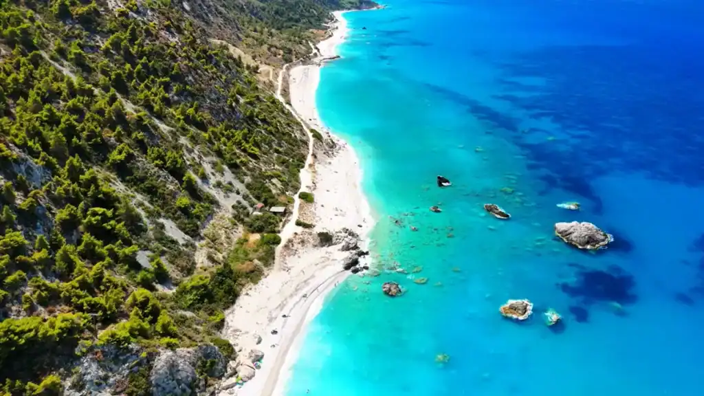 Kathisma Beach stretches along the base of a densely pine-covered hillside, scattered rocks sitting in vivid turquoise shallows that deepen to cobalt blue offshore