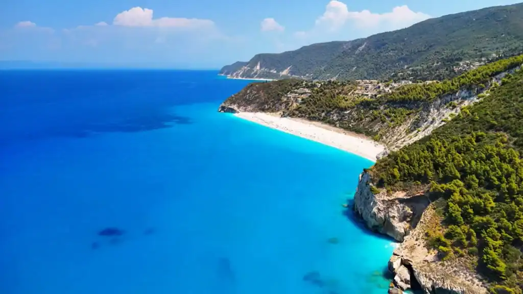 Kathisma Beach seen from above, a pale crescent of sand wedged between dense pine-covered slopes and intensely blue water, with the west coast continuing northward into the distance