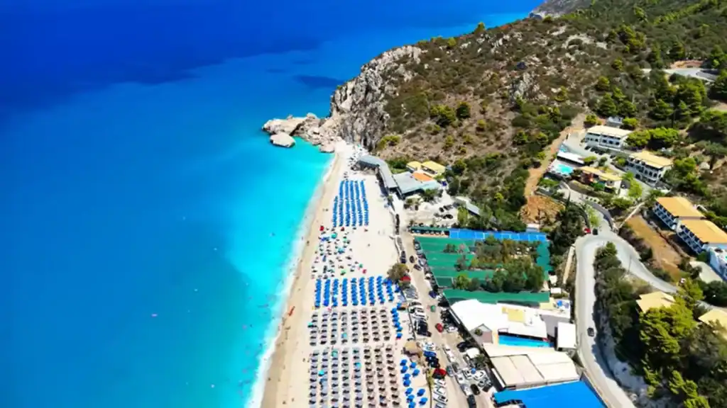 Rows of blue sunbeds packed along a long sandy beach below a rocky scrub-covered hillside, a access road and cluster of buildings tight against the slope to the ri