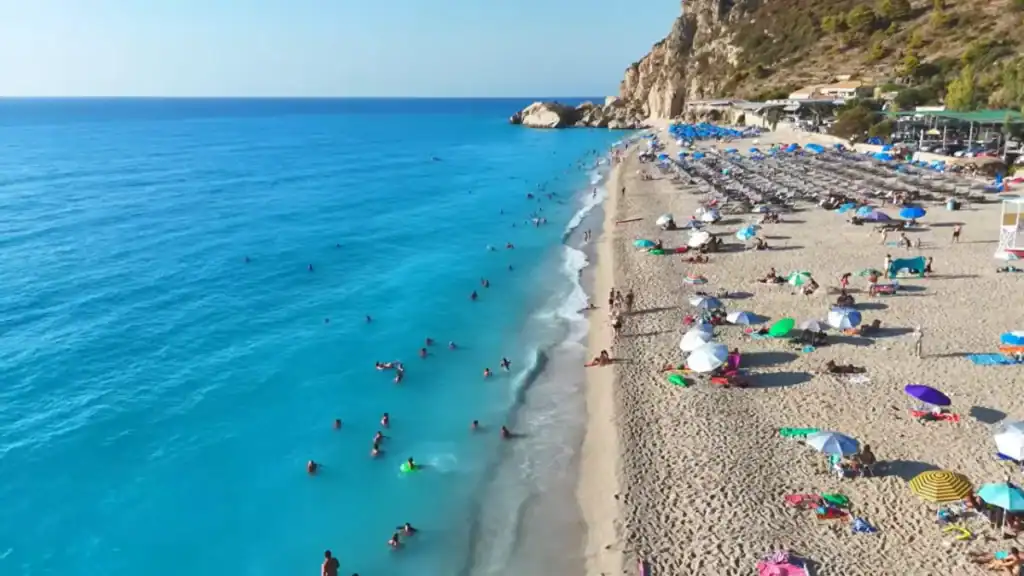 Swimmers spread across the brilliant blue shallows of Kathisma beach while sunbathers rest under scattered umbrellas on the wide pebbly shore, a limestone headland closing off the far end