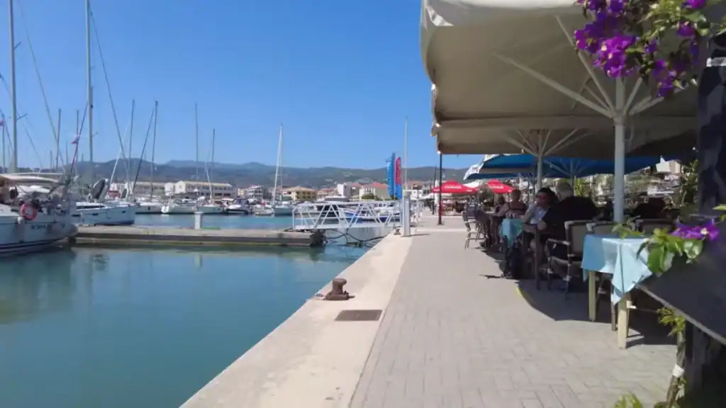 Sailing yachts berthed along a calm marina quay, with diners seated at a waterside restaurant terrace framed by bougainvillea, hills visible across the water.