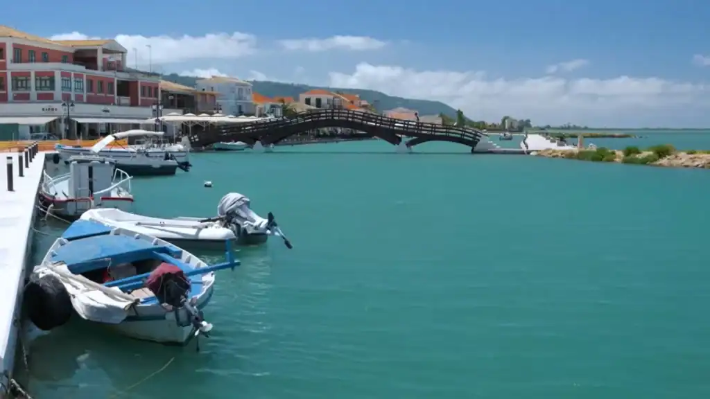 Small boats moored on turquoise harbour water in Lefkada town, with a arched wooden footbridge crossing toward a low breakwater and green hills beyond.