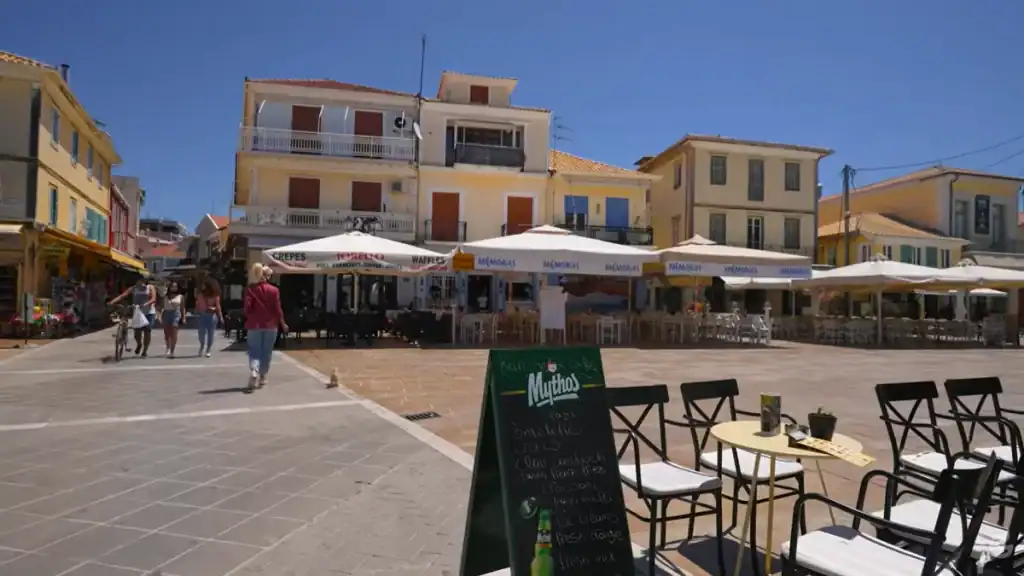 Broad open square with café terraces under white parasols, a Mythos chalkboard menu in the foreground, and pedestrians crossing toward yellow-rendered buildings opposite.