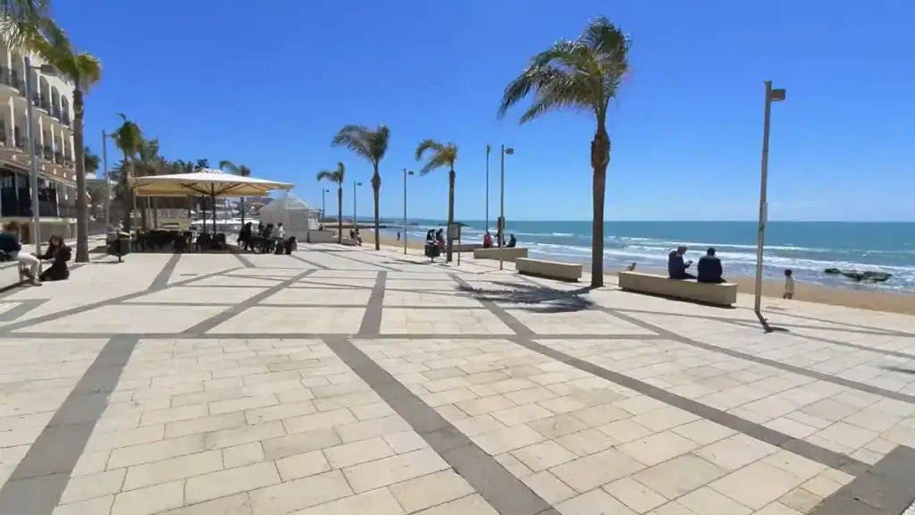 Wide patterned stone esplanade with palm trees, people seated on benches facing the sea, and a café terrace with parasols beside a seafront hotel on the left.