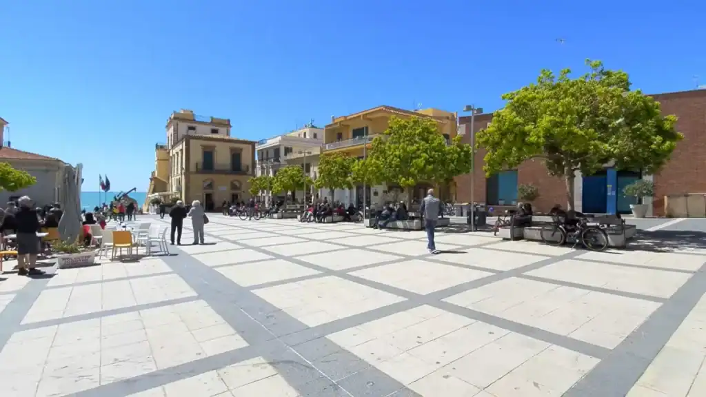Paved town square with people gathered on benches beneath green trees, café tables to one side, and a glimpse of the sea visible between buildings at the far end.