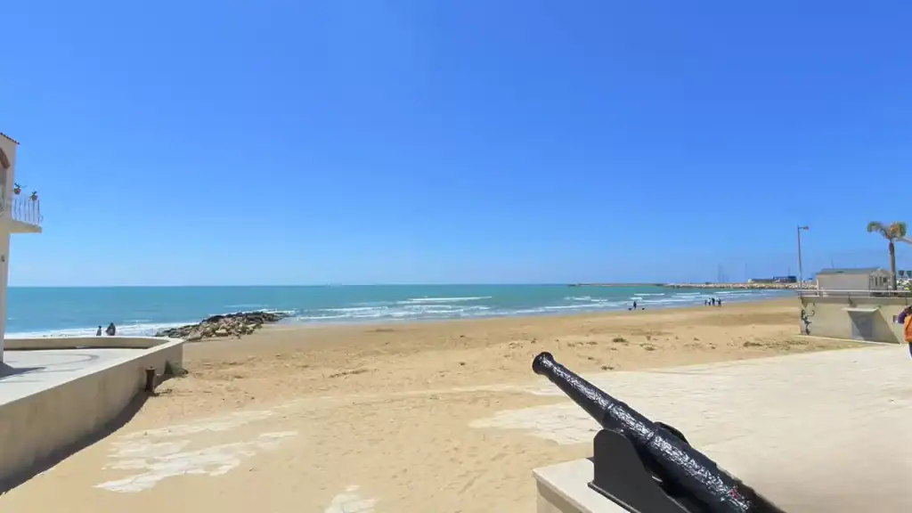Black iron cannon mounted on the promenade wall with a wide sandy beach stretching toward the harbour breakwater, a few figures walking the shoreline in the distance.