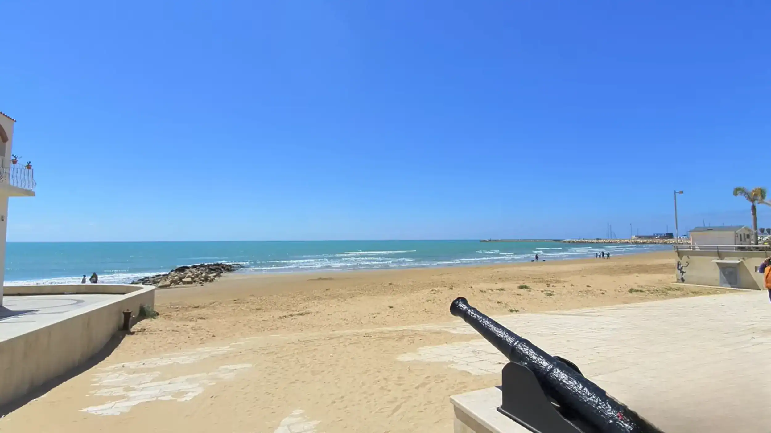 Black iron cannon mounted on the promenade wall with a wide sandy beach stretching toward the harbour breakwater, a few figures walking the shoreline in the distance.