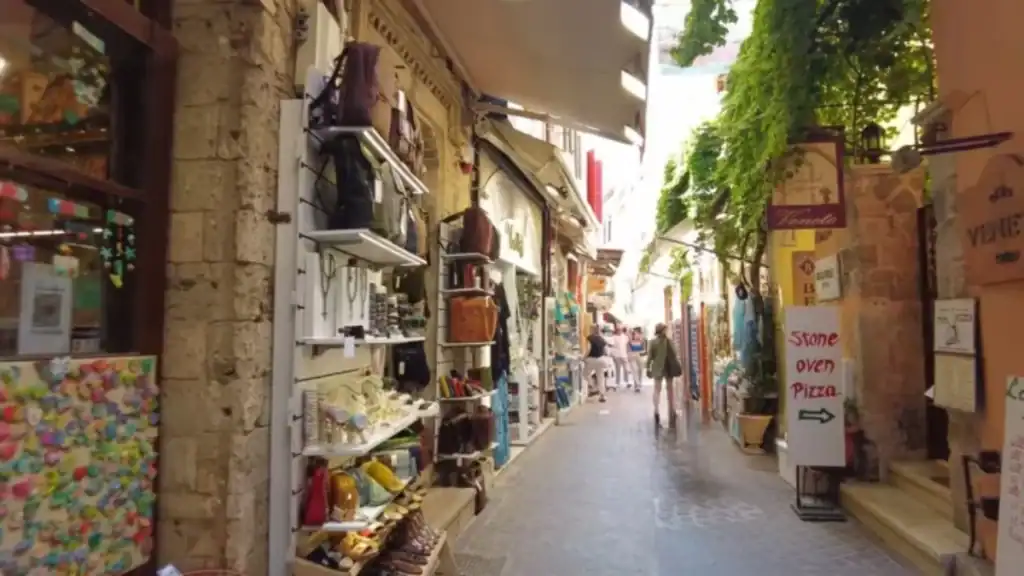 Narrow back street in Chania's old town lined with leather goods, ceramics, and jewellery spilling from shop fronts, a stone oven pizza sign pointing right