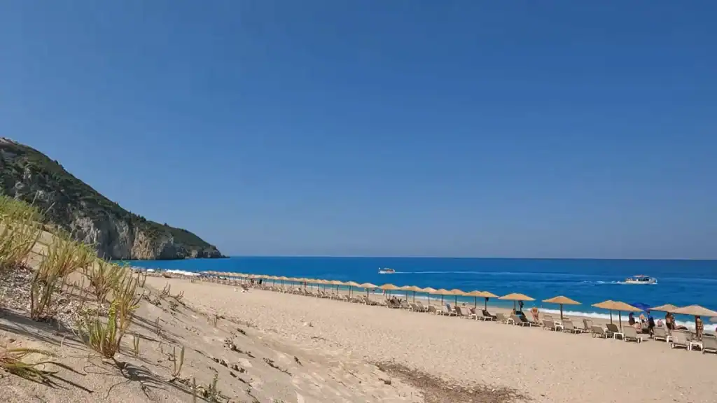 Rows of straw parasols and sunloungers stretching along Milos Beach, with a scrub-covered headland closing the bay to the left and deep blue water beyond