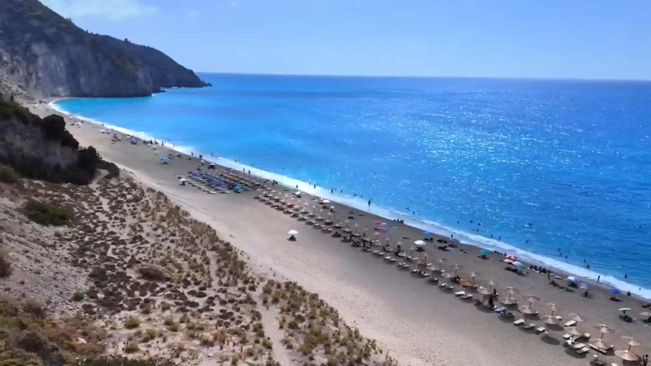 Rows of sun loungers and parasols along Milos Beach, Lefkada, with intense turquoise water and a steep cliff face closing off the northern end.