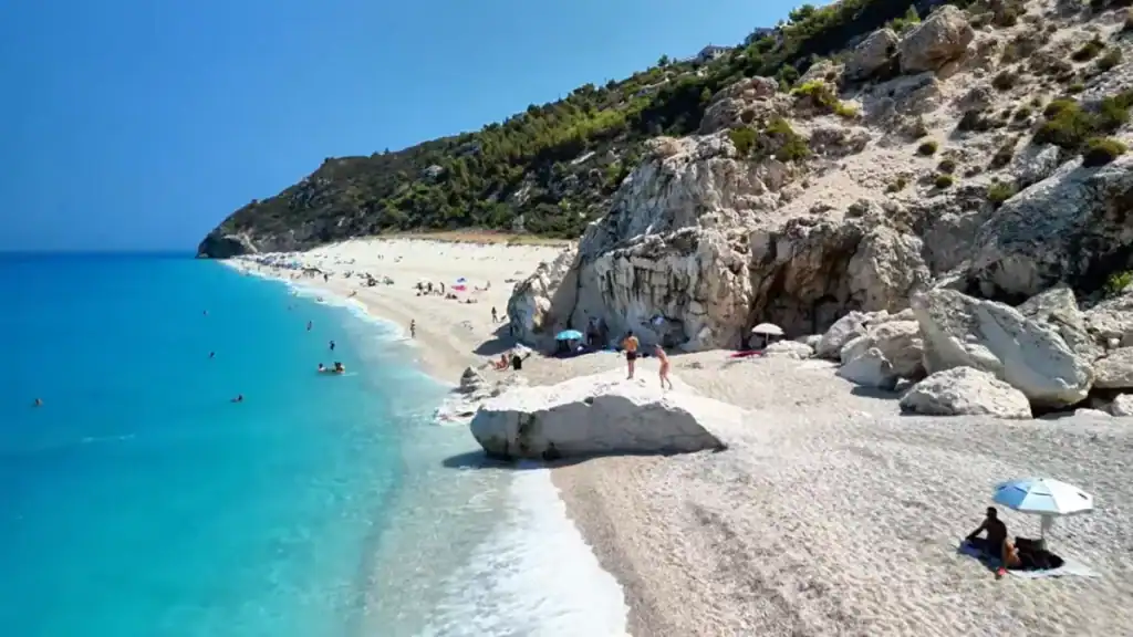 Pale limestone boulders jut onto the shingle at the southern end of the beach, with swimmers spread through shallow turquoise shallows and a wooded cliff rising steeply behind.