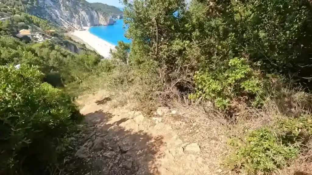 Dry scrub and maquis press in on both sides of a rocky footpath descending toward Milos Beach, visible below as a crescent of white sand against vivid turquoise water.