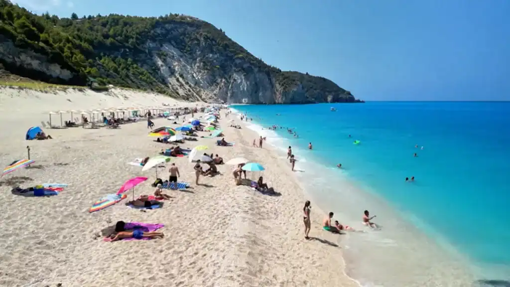 Wide sandy shoreline stretching toward a forested limestone headland, sunbathers and swimmers spread along the water's edge with intense blue sea extending to the right