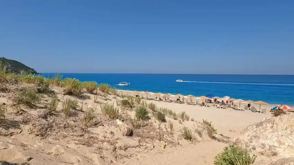 Sparse vegetation covers a sandy rise in the foreground, with thatched parasols and loungers arranged along the waterline and two speedboats cutting across deep blue open water.