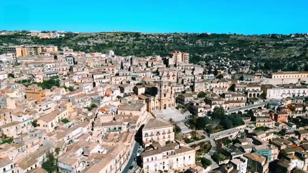 Rooftops of Modica spread across a shallow valley, the Cathedral of San Giorgio's facade prominent at centre, with the town thinning toward scrubland and low hills behind.