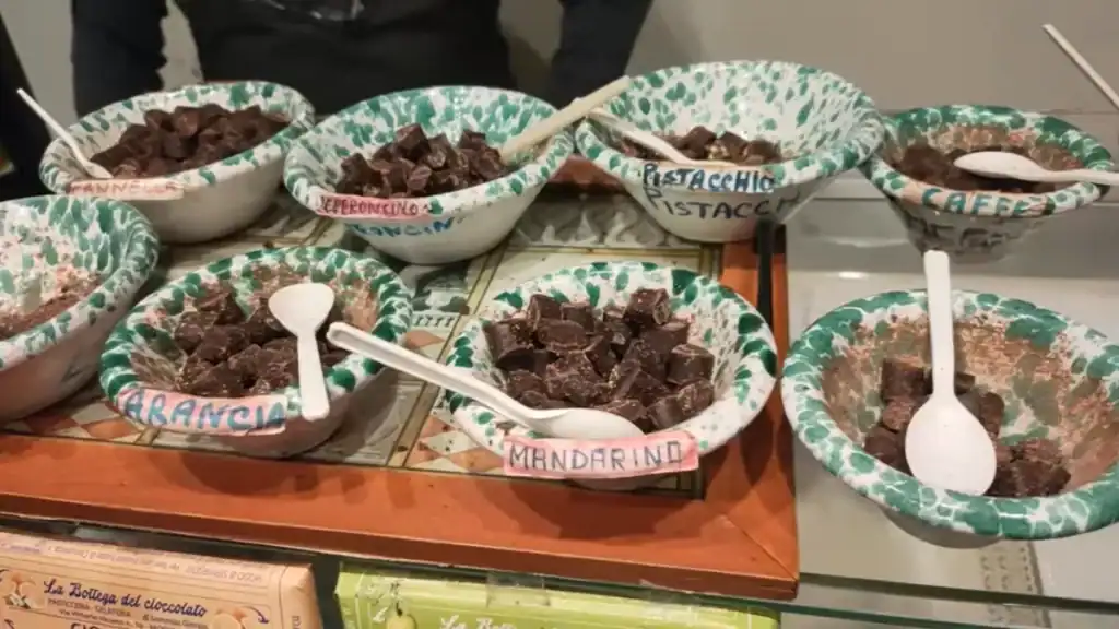 Tasting bowls of Modica chocolate in flavours including mandarin, orange, pistachio, chilli and coffee, served in hand-painted Sicilian ceramic dishes with small white spoons.