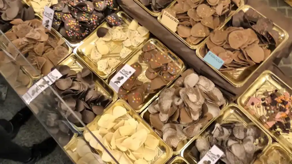Trays of traditional Modica chocolate discs in dark, milk, white and spiced varieties fill a shop counter display, labelled with flavours including zenzero and fragola