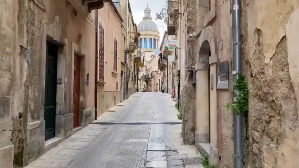Quiet flagstone street in Ragusa Ibla lined with weathered limestone facades, the dome of San Giorgio visible at the far end