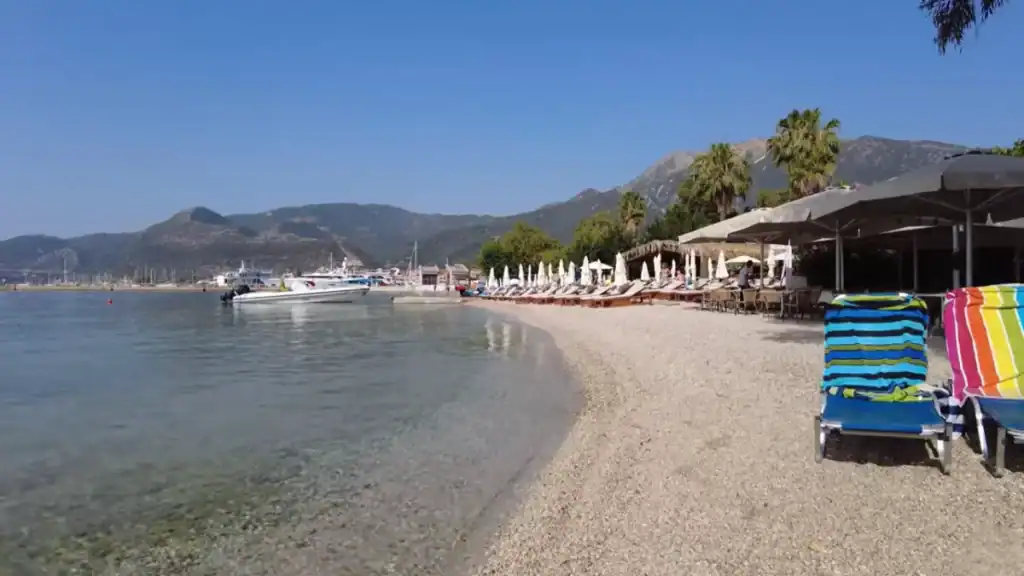 Striped beach towels on sunloungers at the water's edge on Nidri beach, a shingle shoreline curving toward the harbour where speedboats are moored, palm trees and a beach bar behind, steep mountains across the bay