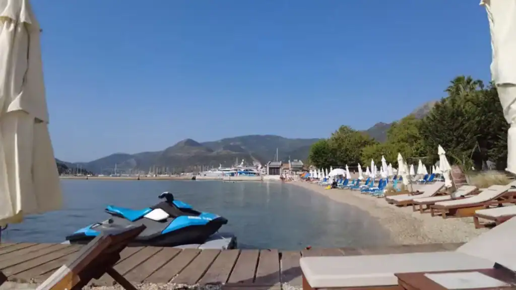 Nidri Lefkada beach with a jet ski on a wooden platform in the foreground, rows of sunloungers and closed parasols along the shoreline, and the harbour with moored yachts visible further along the bay