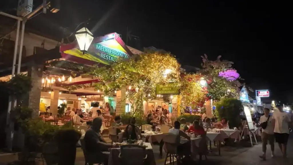 Busy outdoor restaurant terrace on Nidri's main strip at night, with diners at checked tablecloth tables beneath fairy-lit trees and a sign for Panorama Greek Restaurant visible among the warm glow