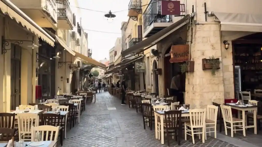Outdoor restaurant tables and chairs line a narrow cobblestone alley in Chania Old Town, with stone-built facades and canvas awnings overhead