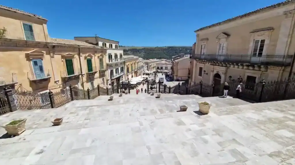 Broad stone steps descending from a church forecourt toward the main street of Ragusa Ibla, Baroque palazzi with wrought-iron balconies on both sides and a piazza with café umbrellas below.