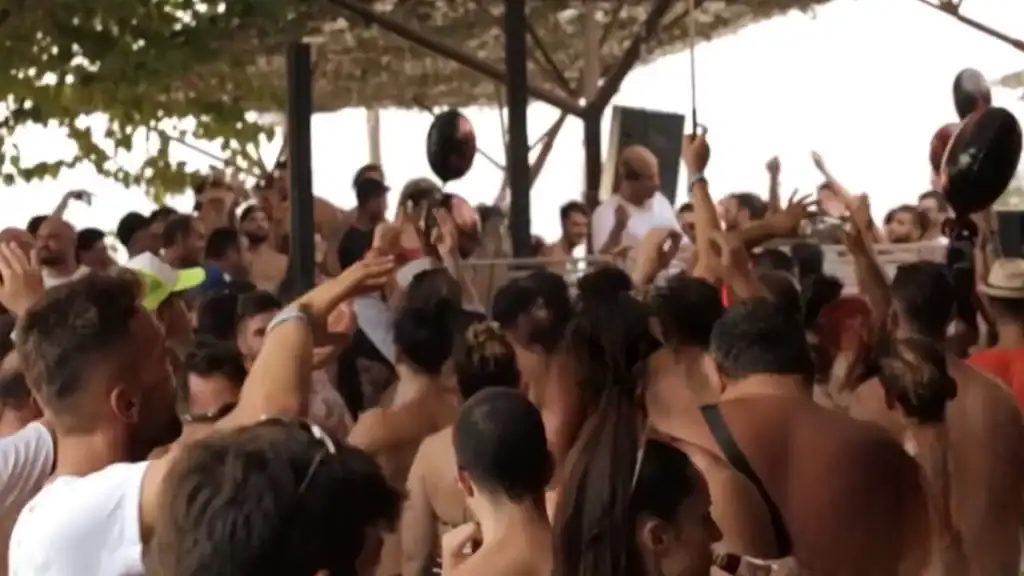 Packed crowd dancing under a thatched canopy at Kathisma beach, arms raised toward a DJ on a low stage, black balloons drifting above the throng