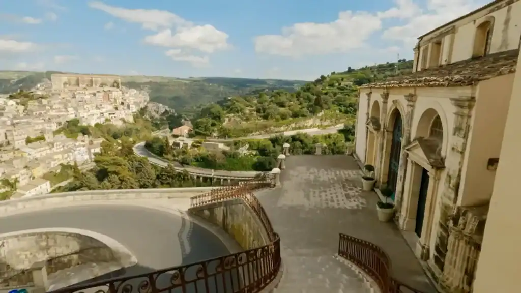 Stone terrace beside a Baroque church facade, with a curved iron balustrade and a hillside town spreading across the valley below.