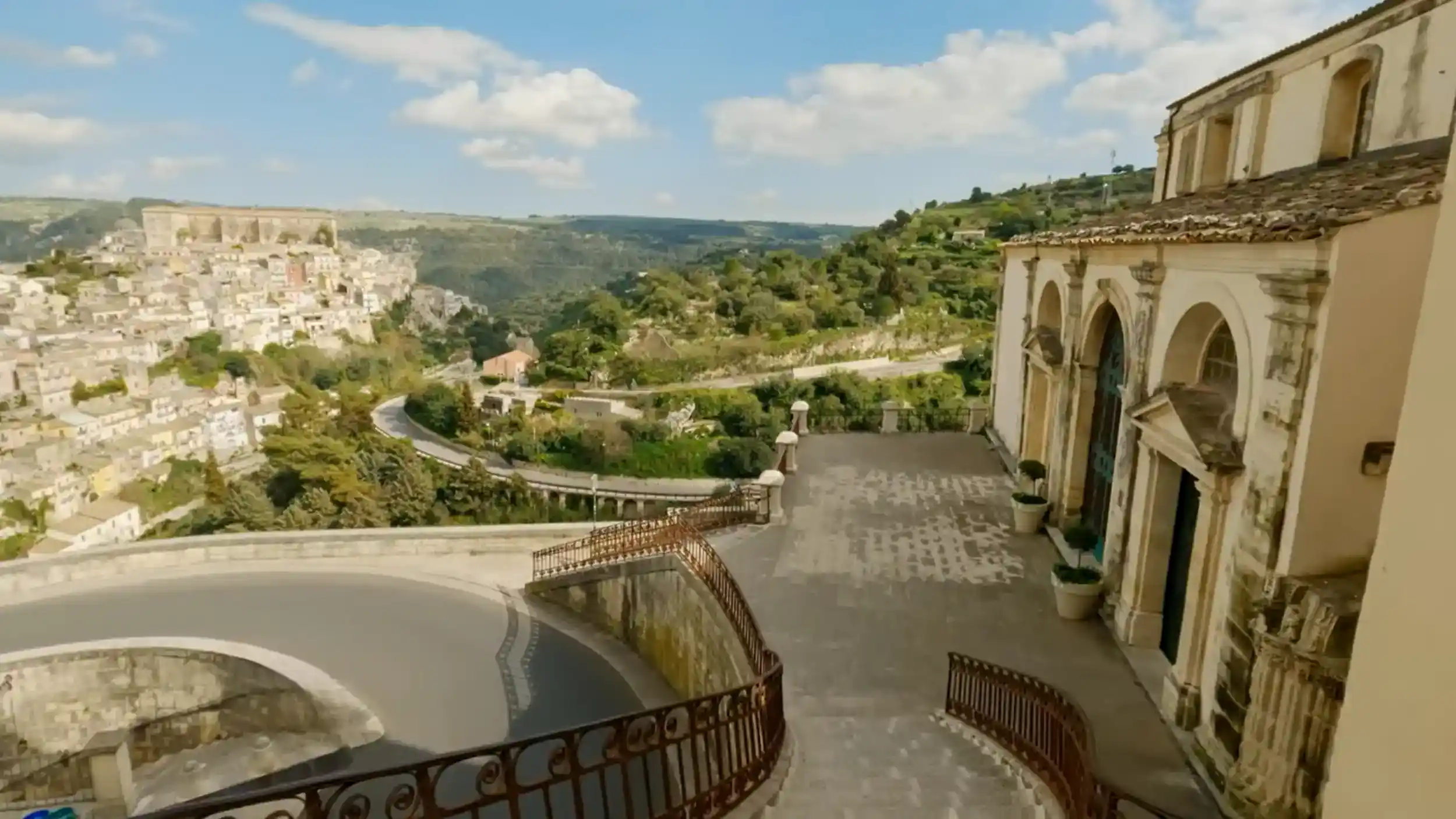 Stone terrace beside a Baroque church facade, with a curved iron balustrade and a hillside town spreading across the valley below.