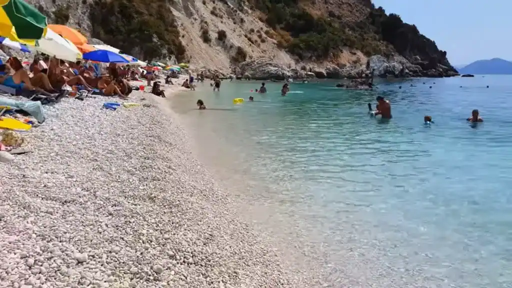 Smooth white pebbles cover the shoreline at Agiofili Beach where swimmers wade in shallow clear water, colourful umbrellas crowding the upper shore beneath rocky cliffs