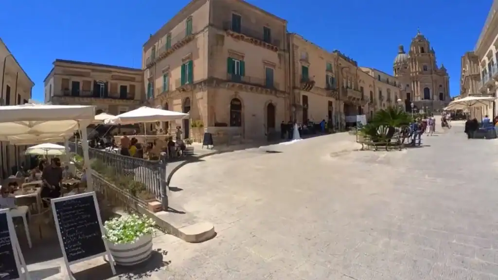 Busy piazza in Ragusa Ibla with outdoor café seating, pale stone buildings, and the dome of the Cattedrale di San Giorgio rising at the far end.