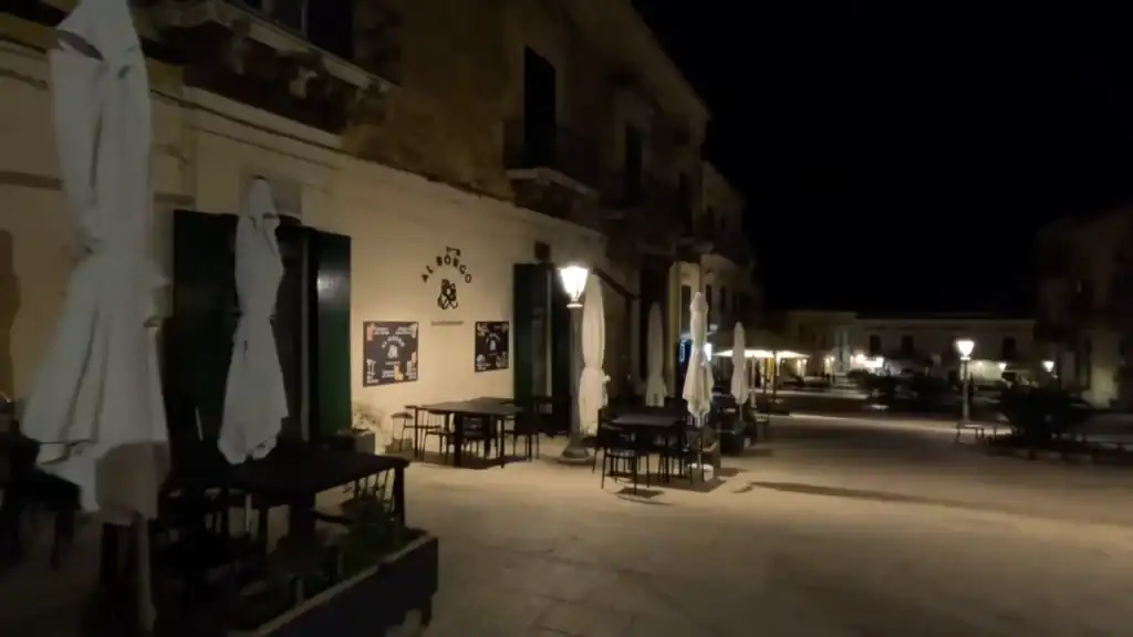 Empty café terrace on a quiet piazza in Ragusa Ibla at night, street lamps casting warm pools across the stone paving