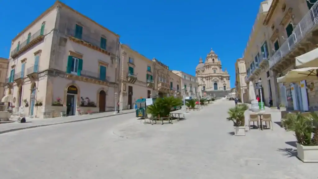 Piazza Duomo in Ragusa Ibla stretching toward the twin-towered facade of the Duomo di San Giorgio, palm trees and cafe tables along the edges - walking in Ragusa Ibla ends here for many visitors