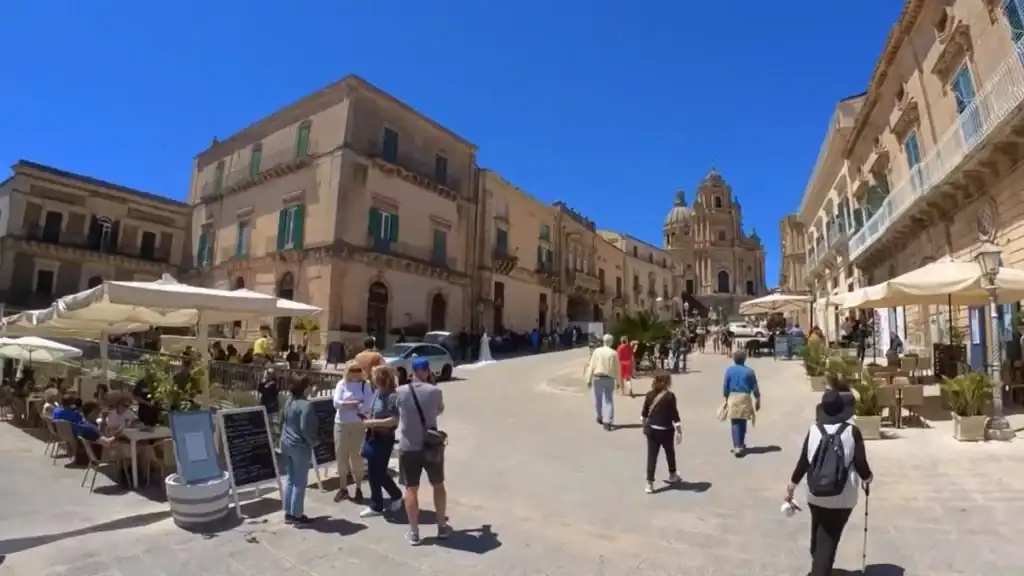 Piazza Duomo in Ragusa Ibla on a busy midday, pedestrians and outdoor cafe seating filling the square with the Cathedral of San Giorgio's dome visible at the far end - the kind of scene that defines travelling between Modica and Ragusa.