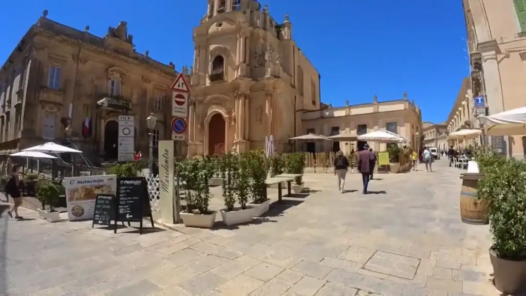 Pedestrians crossing a sunlit paved square in Ragusa Superiore, cafe seating and potted plants in the foreground with the Baroque facade of the Cathedral of San Giovanni Battista behind.