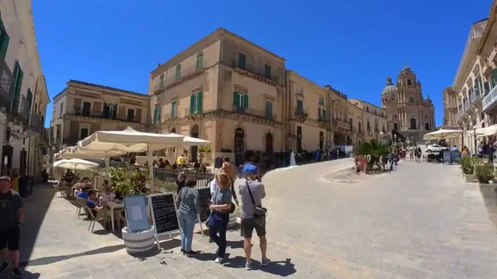 Busy stone-paved square in Ragusa Ibla with café tables to the left, visitors gathered near a menu board, and the dome of the Cathedral of San Giorgio visible at the far right.