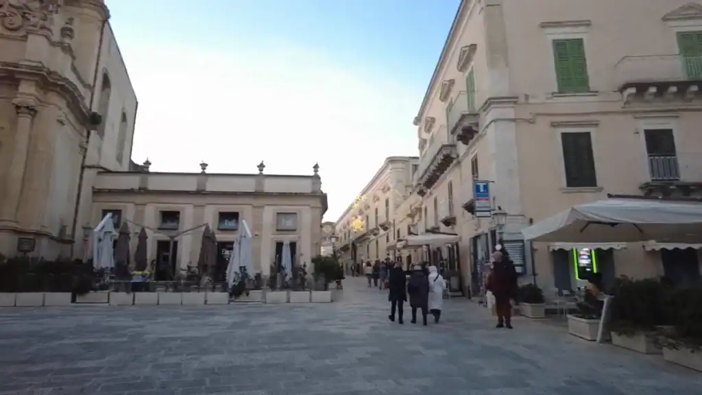 Pedestrians crossing Piazza Pola in Ragusa Superiore at dusk, with the Church of San Giuseppe on the left and the main corso stretching away between pale limestone palazzi.