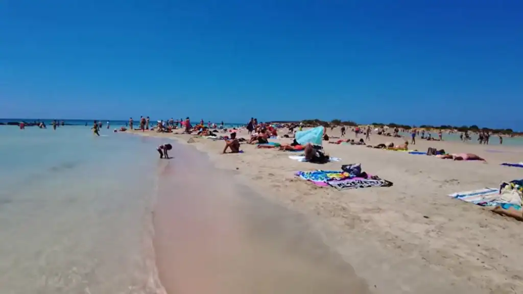 Crowds of sunbathers spread across a wide pale pink sandy beach beside clear shallow turquoise water under an cloudless blue sky