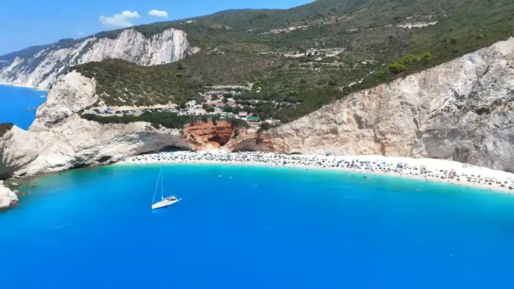 Porto Katsiki's crowded white shoreline tucked beneath forested cliffs, a sailboat drifting across intensely blue water in the foreground