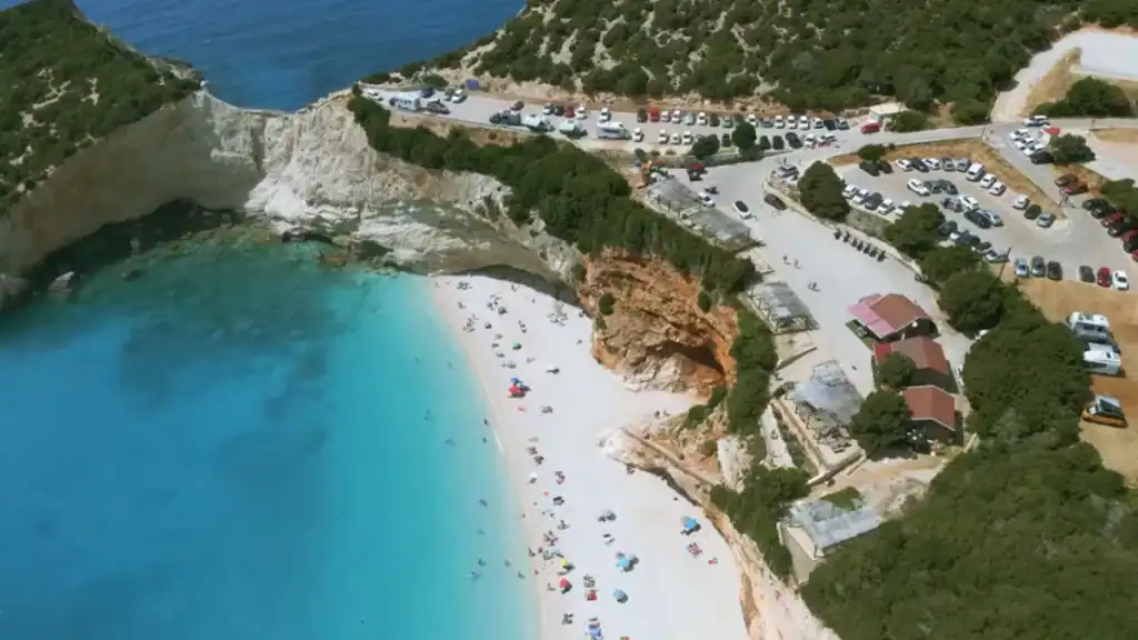 Porto Katsiki beach from above, its white sand and turquoise water flanked by orange and white cliffs, with a busy car park and facilities visible on the plateau above.
