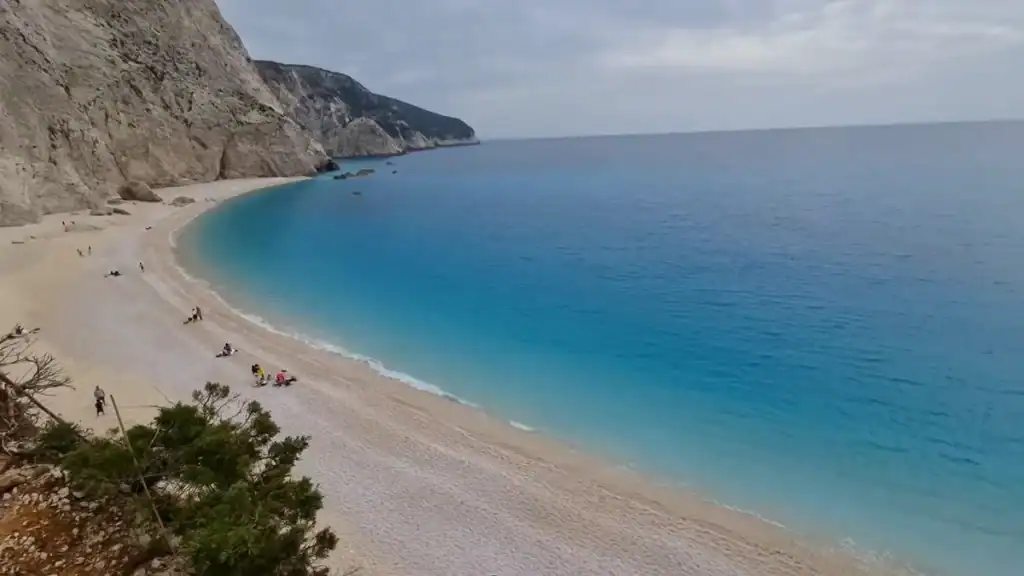 Porto Katsiki beach curves beneath sheer white limestone cliffs, its turquoise water meeting a wide shingle shoreline with a handful of visitors scattered along it
