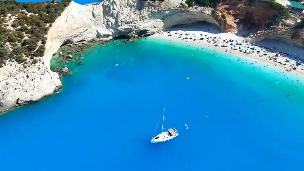 White limestone cliffs curving around Porto Katsiki's turquoise bay, a sailboat anchored in deep blue water below. One of Lefkada's best beaches.