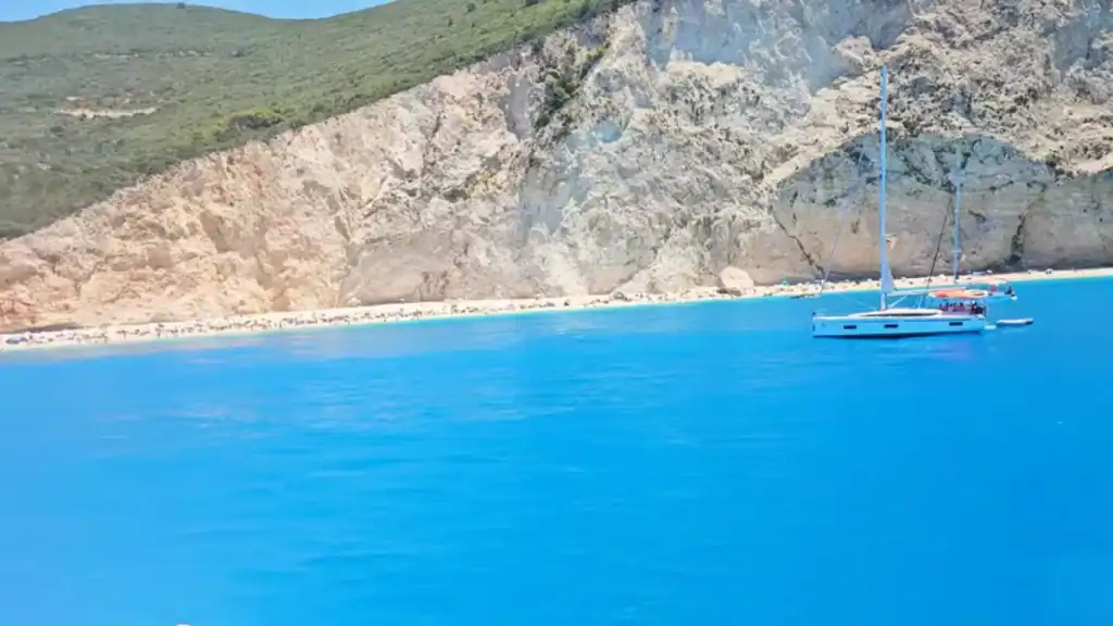Saturated turquoise water filling the foreground with a sailboat anchored offshore, beachgoers spread along a narrow strip of sand backed by fractured limestone cliffs and forested slopes.
