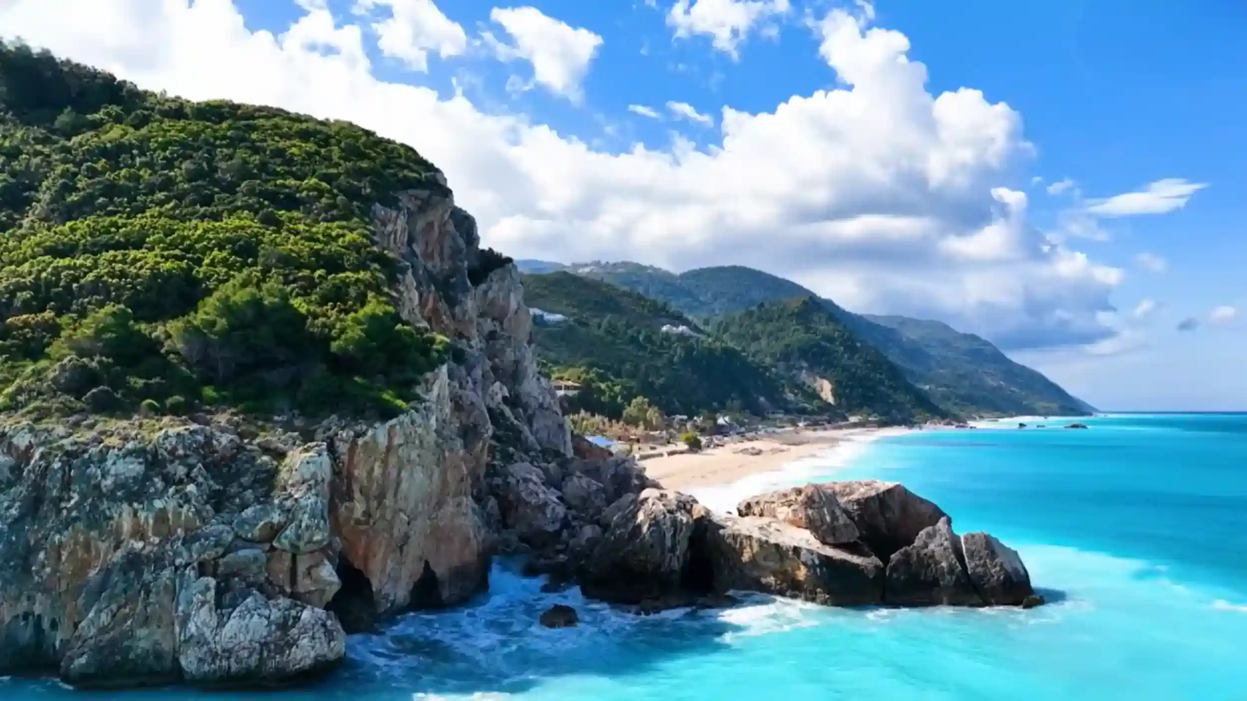 Porto Katsiki beach stretches behind foreground cliffs of weathered rock, the water an intense turquoise against the pale sand, forested hills running along the coastline behind.