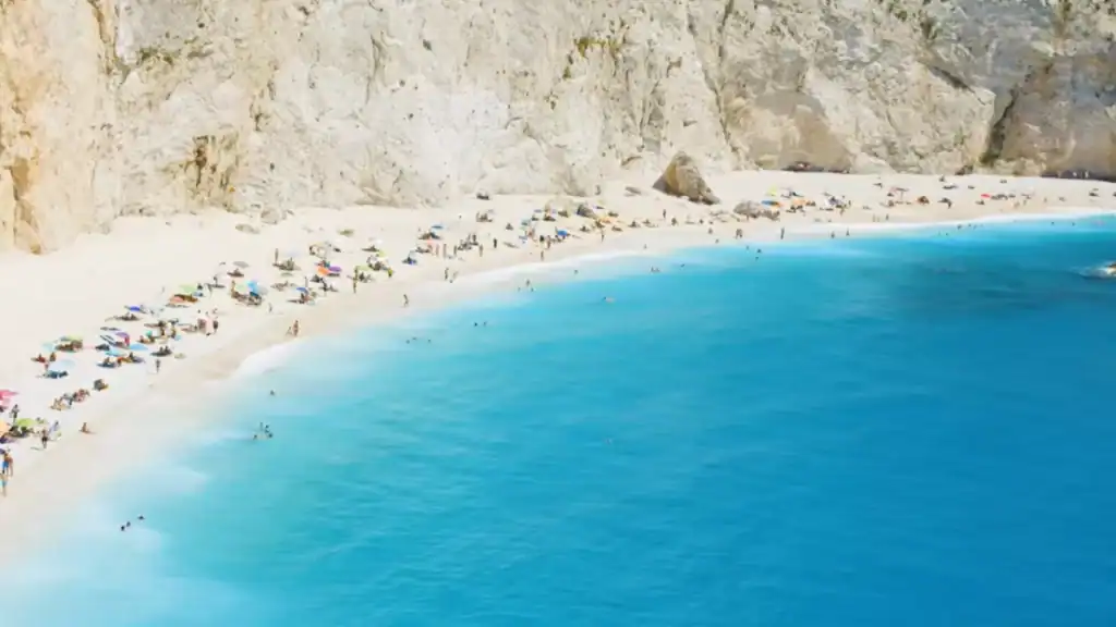 Crowded white sand beach dotted with colourful umbrellas, backed by sheer pale limestone cliffs, swimmers scattered across vivid blue water in the foreground.